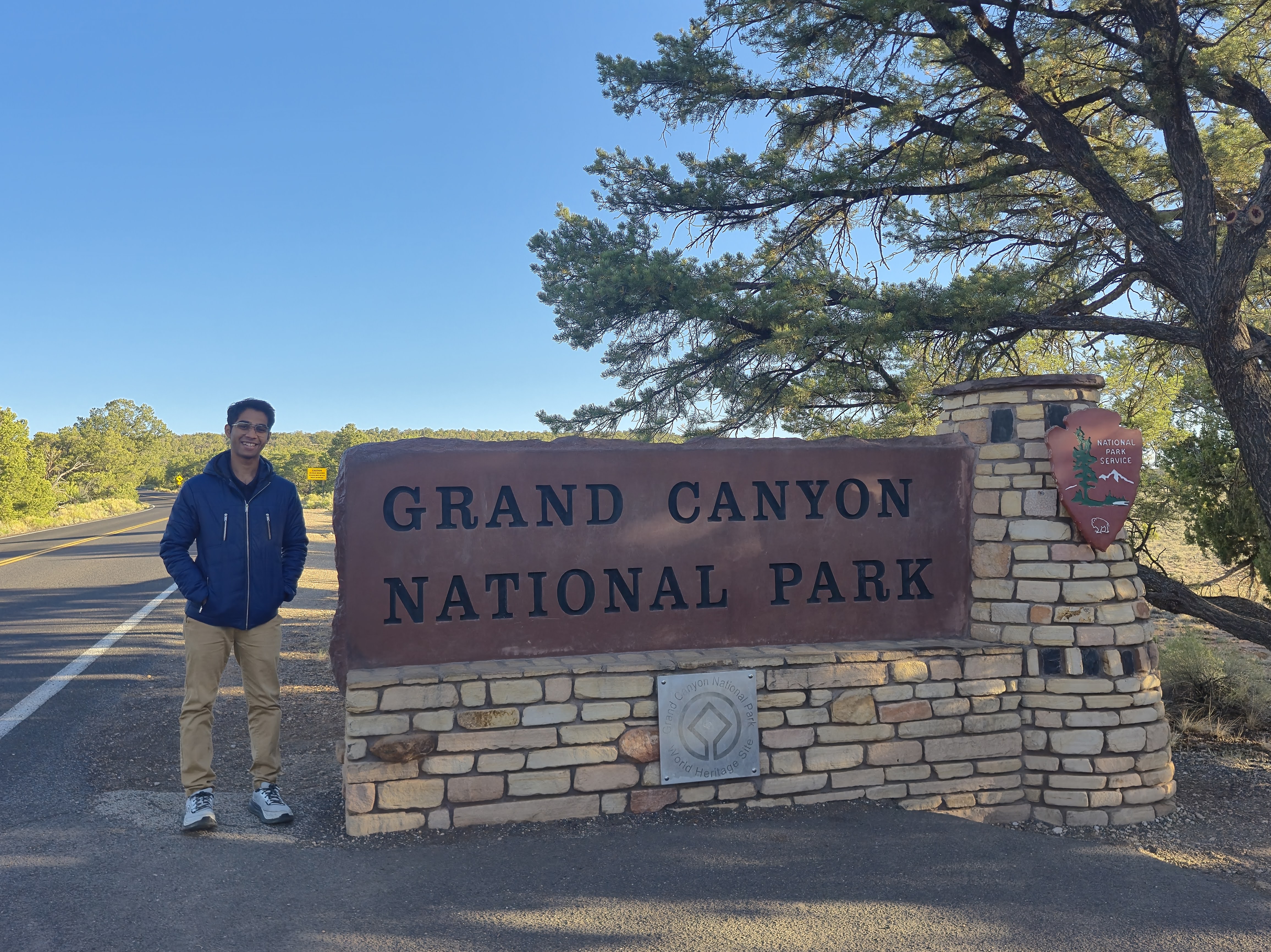 Aldrin standing beside the stone 'Grand Canyon National Park' entrance sign on a sunny morning
