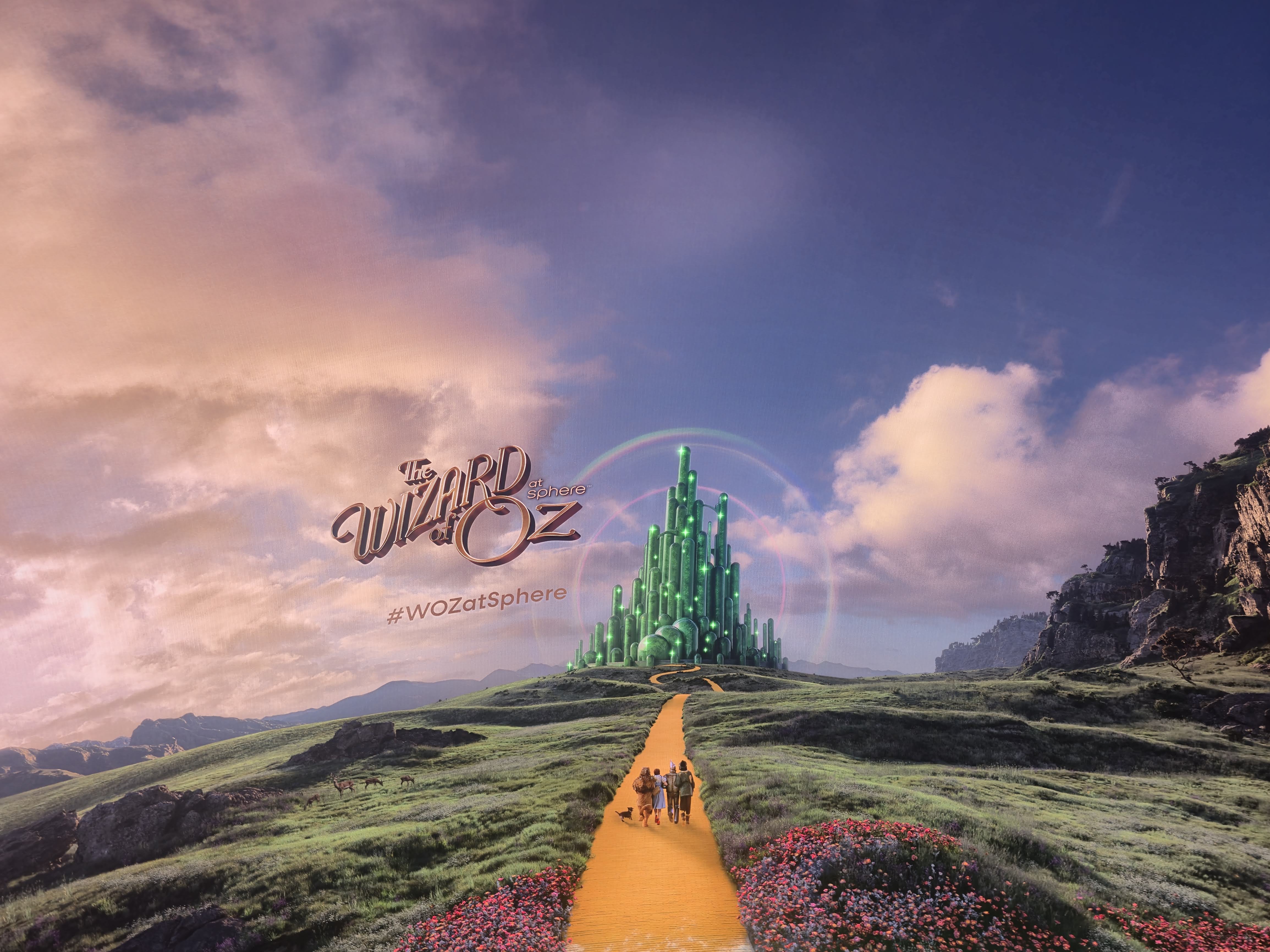 Promotional artwork for 'The Wizard of Oz at Sphere' — the Emerald City at the end of a yellow brick road winding through green hills and a field of red poppies