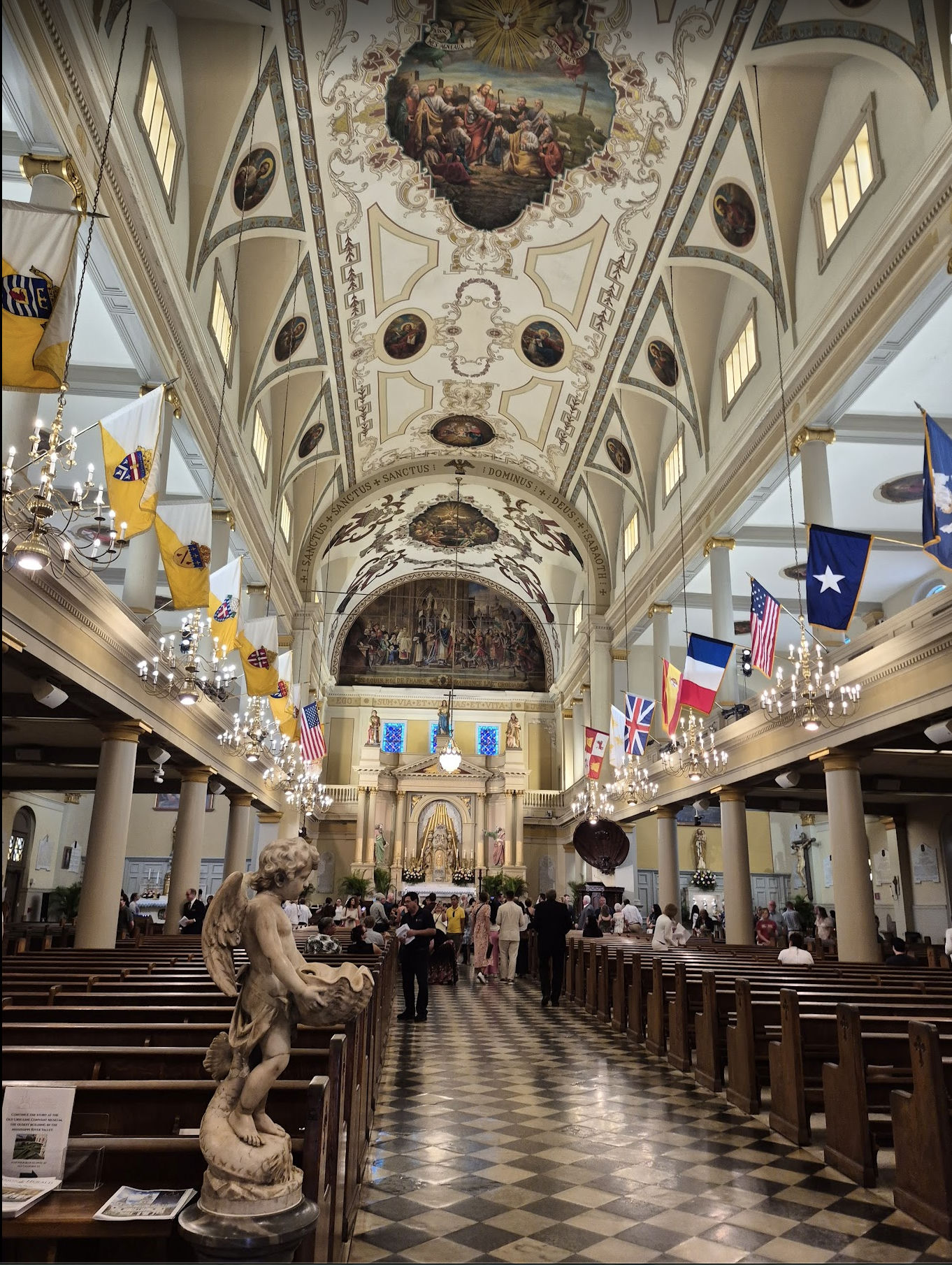 The ornate interior of St. Louis Cathedral during Mass — a vaulted, painted ceiling, chandeliers, flags hanging in the nave, and a black-and-white checkerboard marble floor